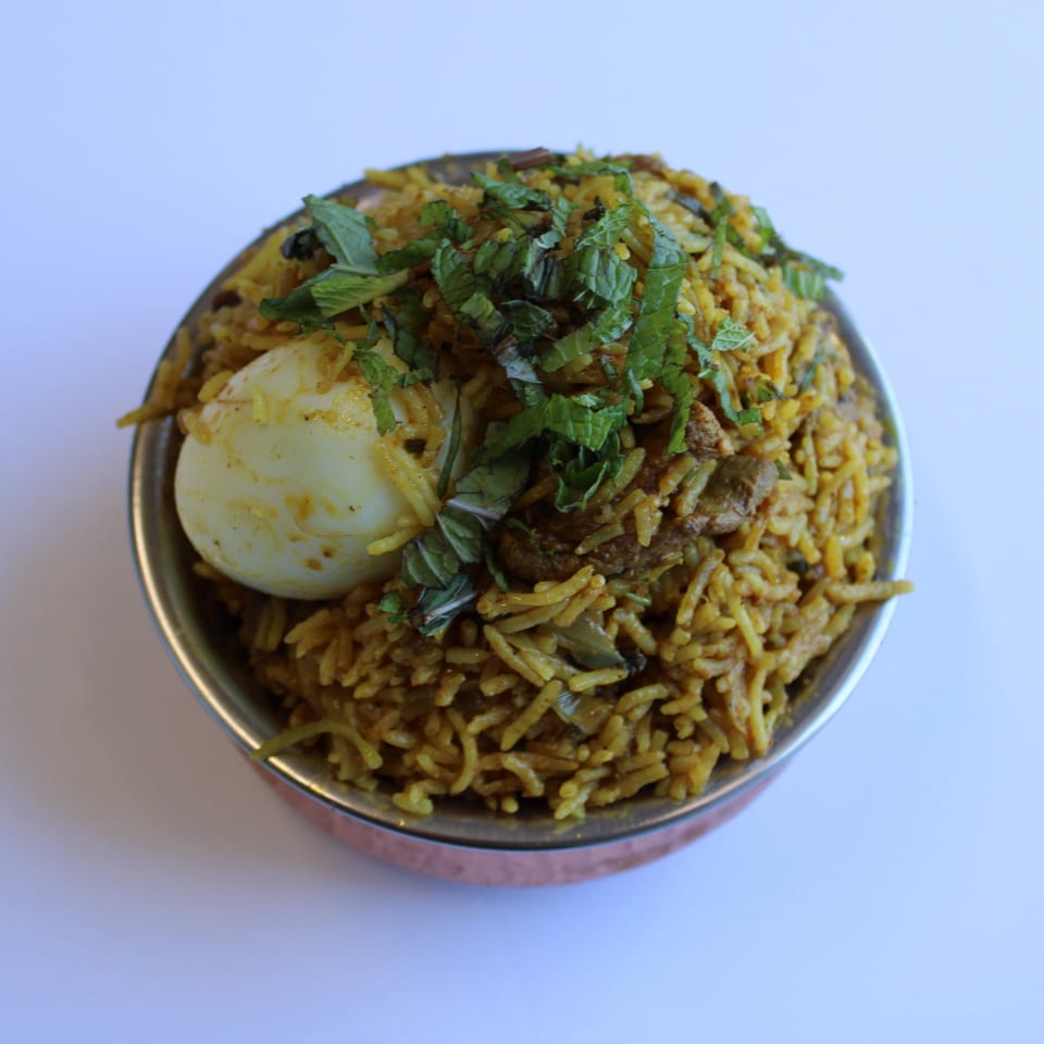 Indian biryani with rice, meat, a hard-boiled egg, and fresh mint in a copper bowl.