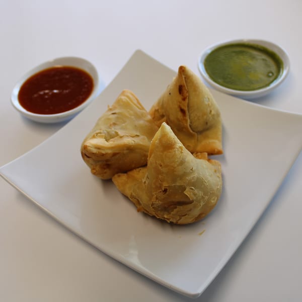 Three golden-brown samosas on a white plate, with red and green chutneys in small bowls.
